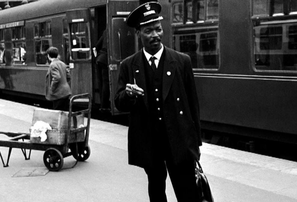 A black and white old looking photo of a person called Asquith Xavier standing in front of a train wearing a rail uniform