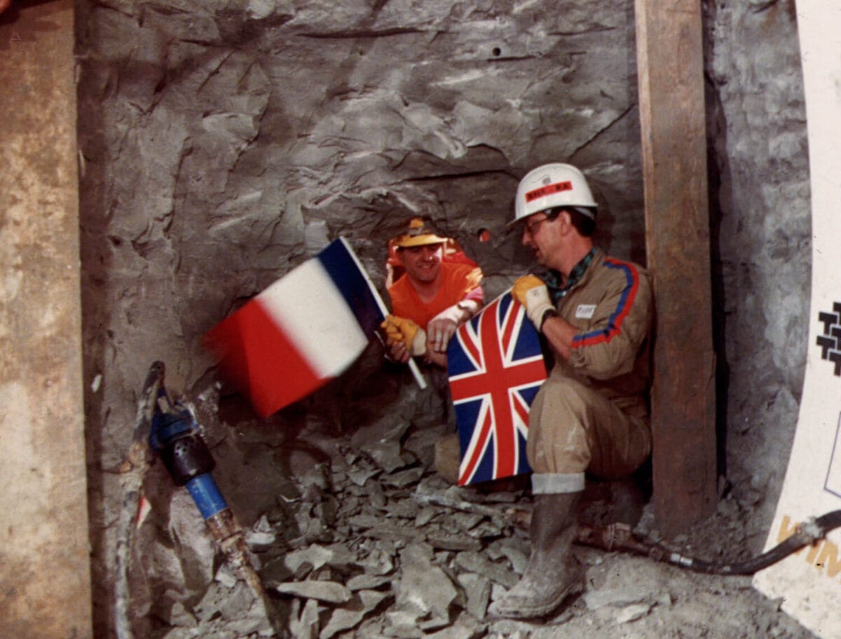 Two people reaching through a whole in the channel tunnel, one holding a UK flag and one holding a French flag