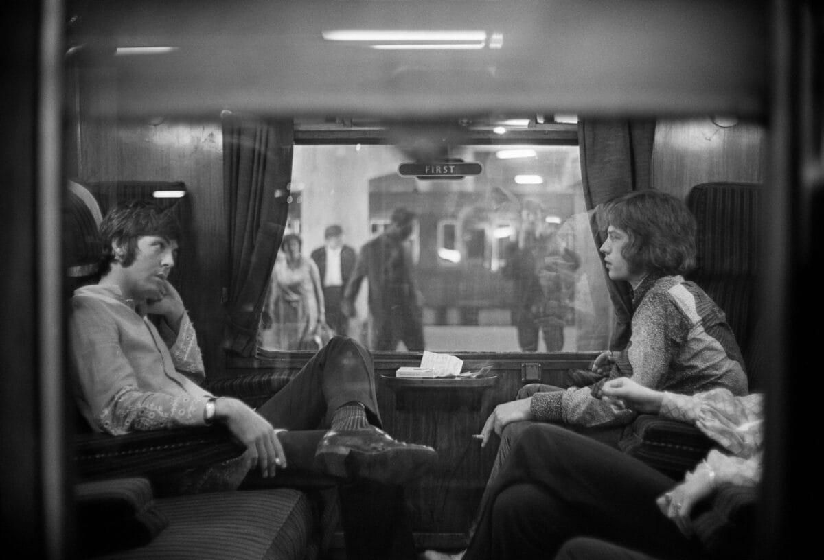 An old photo of Paul McCartney of the Beatles and Mick Jagger of the Rolling Stones sat opposite each other on a train at Euston Station