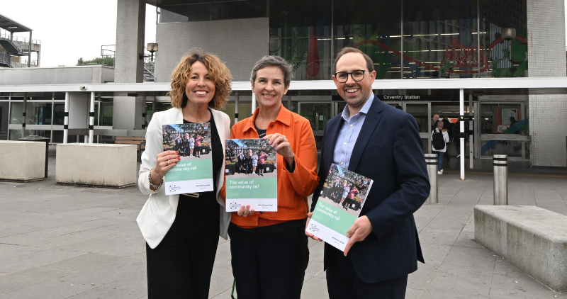 Two women and a man standing in front of Coventry train station holding the Value of Community Rail report