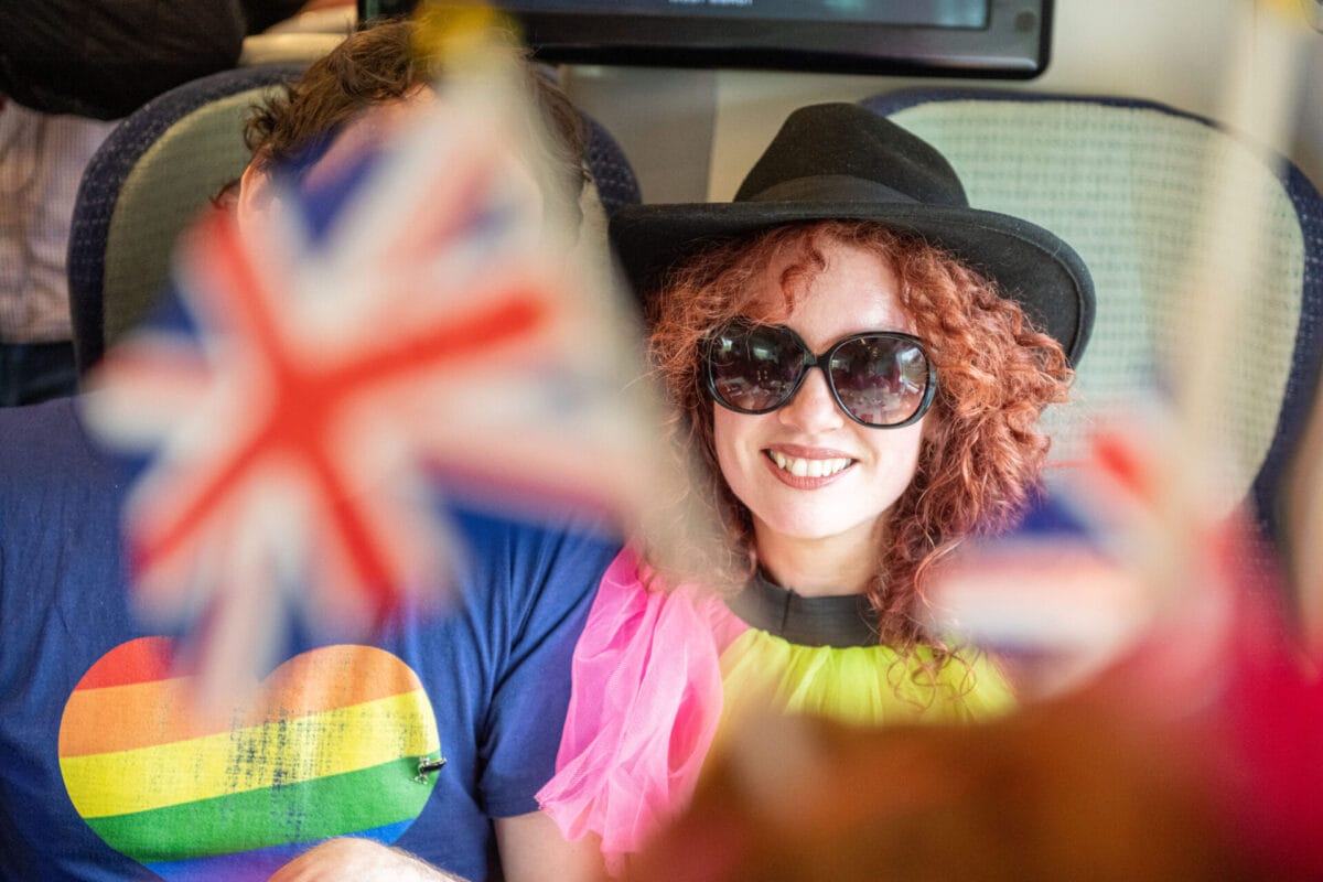 A woman sitting on a train smiling wearing sunglasses waving the Union flag celebrating the Eurovision song contest