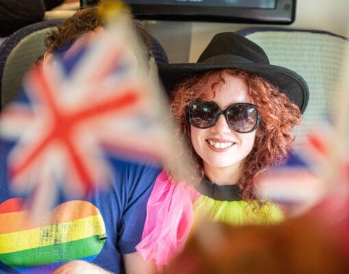 A woman sitting on a train smiling wearing sunglasses waving the Union flag celebrating the Eurovision song contest
