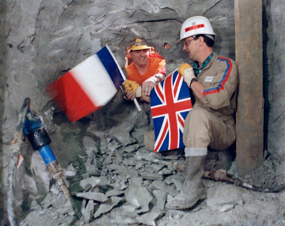 Two people reaching through a whole in the channel tunnel, one holding a UK flag and one holding a French flag