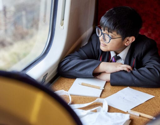 A school boy sitting in a train looking out of the window with a pencil and paper on the table in front of him.