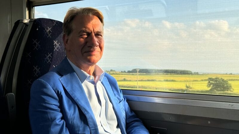 A man on a train, smiling. Green fields are visible through the window.