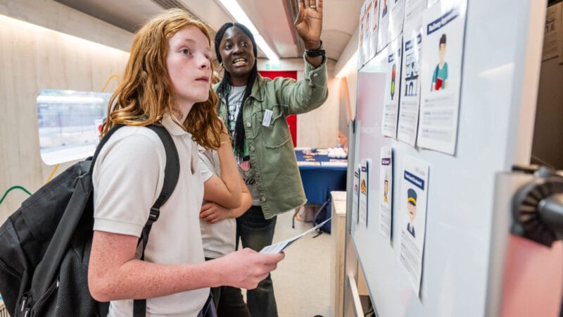 A young person in school uniform looks at a whiteboard full of careers as an adult points out roles.