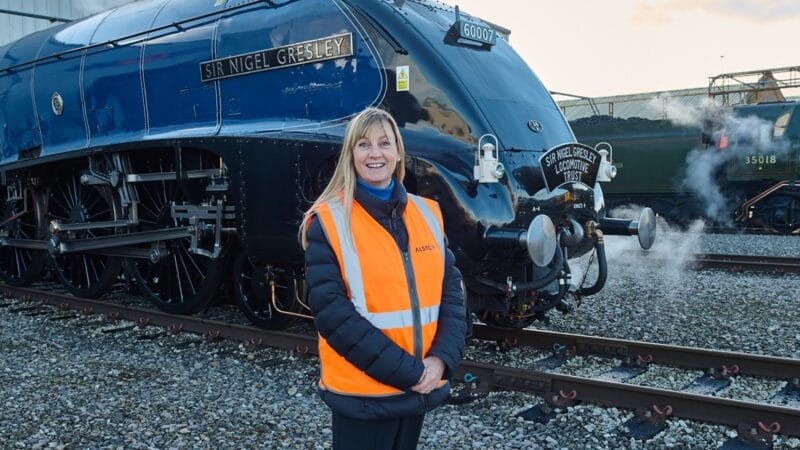 Alstom Communications Manager Kathryn Lancaster stands in front of the locomotive 'Sir Nigel Gresley'. She is wearing orange high-vis and steam is coming from the blue locomotive